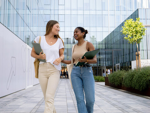 Two diverse teenage female students with folders walking and chatting in a campus - Powered by Adobe