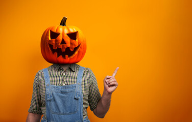 Person in Halloween costume of scarecrow with Jack-o'-lantern pumpkin head points away over orange background with copy space - obrazy, fototapety, plakaty