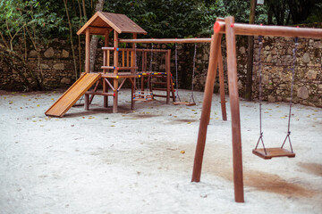 A playground in a tropical setting with a selective focus on a small wooden house with a triangle roof and children's slide; several swings for kids on the sand, ones in a defocused foreground