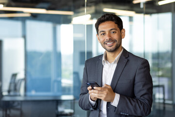 Portrait of young hispanic man inside office at workplace, businessman smiling and looking at...