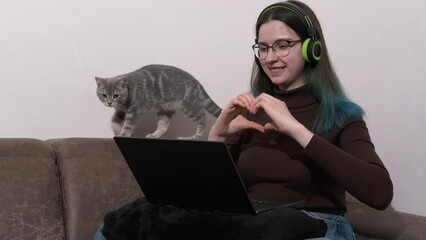 A teenage girl wearing headphones using a laptop, listening to music, communicating online at home. girl shows the shape of heart with her hands, greets friends online. Virtual love. Virtual reality.