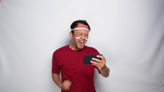 A young Asian man with a happy successful expression wearing red top and headband while streaming on his phone, isolated by white background. Indonesia's independence day concept.