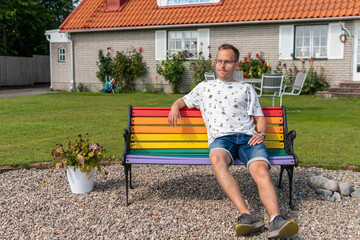 A man sitting outdoors on colorful LGBT+ rainbow bench in front of a private house