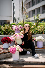 Fototapeta premium Cute young girl surrounded with amazing bouquets with fresh flowers arrangement in hat boxes on blurred street background