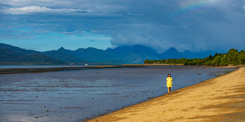pretty girl in yellow dress admiring  the view of hinchinbrook island from the beach in cardwell, north queensland, australia; tropical beach with palm trees and famous island in the background