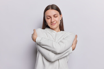 Calm brunette young woman with closed eyes embraces herself wears casual hoodie eprrsses self love...