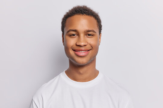 Portrait of pleased dark skinned man with short curly hair stands satisfied glad to hear good news dressed in casual t shirt isolated over white background. People and positive emotions concept