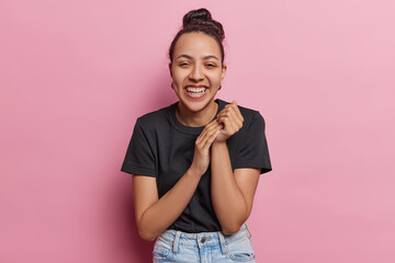 Happy overjoyed Latin woman clasps hands laughs positively smiles broadly shows white teeth dressed in casual black t shirt and jeans isolated over pink background. People and happiness concept