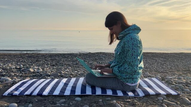 A woman working at a laptop on the seashore. A pretty lady typing on a computer by the sea makes a business transaction online at a distance. Freelance, remote work on vacation.High quality 4k footage