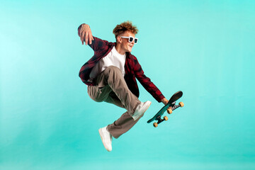 Obraz premium young crazy guy rides skateboard and jumps on blue isolated background, hipster in sunglasses flies