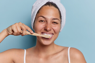 Daily dental care concept. Positive young European woman brushes teeth with wooden toothbrush enjoys morning time wears white soft bath towel on head and t shirt isolated over blue background