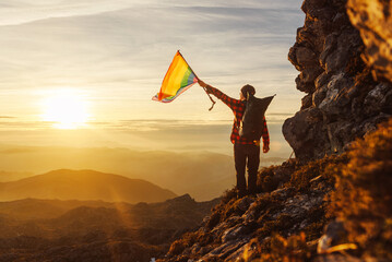 Mountaineer man with backpack on his back waving a rainbow lgbt pride flag at sunset on the mountain .Sexual diversity. Sport and outdoor adventure - obrazy, fototapety, plakaty