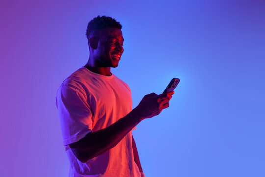 african american man is using smartphone and smiling in neon lighting, guy is typing on phone online
