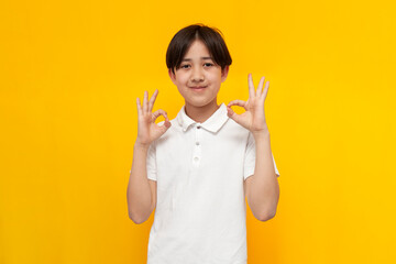 asian boy of twelve years old in white t-shirt shows gesture of ok on yellow isolated background