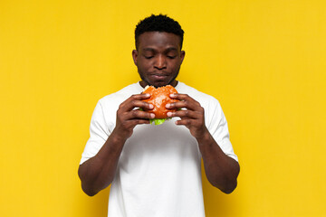 african american man in white t-shirt holding big burger and biting it, the guy eats fast food with his mouth open