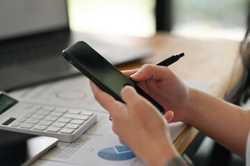 Fototapeta premium Close up with female hand holing smart phone and touching on device screen while doing a financial documents on digital tablet, business data analysis concept.