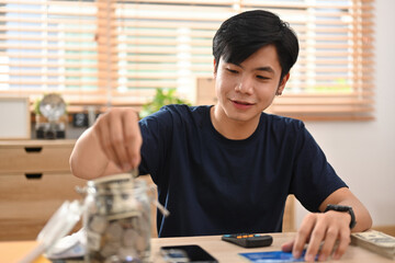 Young Asian man putting Thai currency coin in saving jar to save money purpose for investment or...