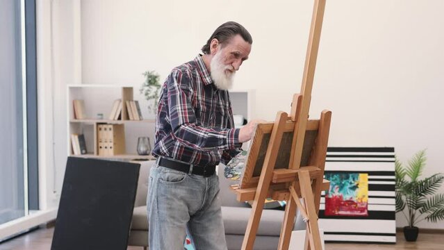 Close up view of elderly male in denim wear with palette layering paint on cloth with brush in studio interior. Involved person honing professional skills and work in designated area of art space.