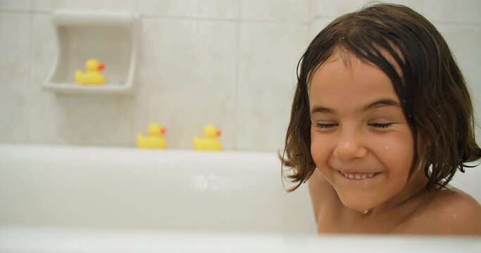 The child splashes with water and is happy while taking a bath. A cheerful smiling girl is bathing in the bathroom. High quality 4k footage