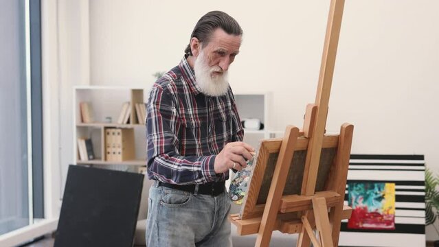 Portrait of confident bearded pensioner with brush posing for camera during creative process in studio. Efficient painter playing freely with ideas, marks, colors and surfaces for success of artwork.