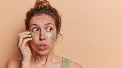 Fototapeta premium Photo of surprised young woman applies refreshing green hydrogel patches beneath her tired eyes to soothe puffiness after sleepless night poses against brown background copy space for your advert