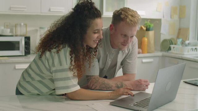 Medium shot of young couple leaning on kitchen table, reading receipt and using laptop while calculating expenses and planning family budget at home