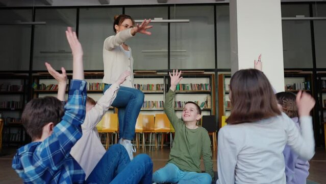 Smiling teacher and kids puts their palms together sitting in circle on floor in library. Concept of teambuilding