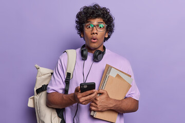 Impressed curly haired Hindu student holding books and notepads carrying backpack uses mobile phone for chatting with friends dressed in tshirt isolated on purple. Education school college concept