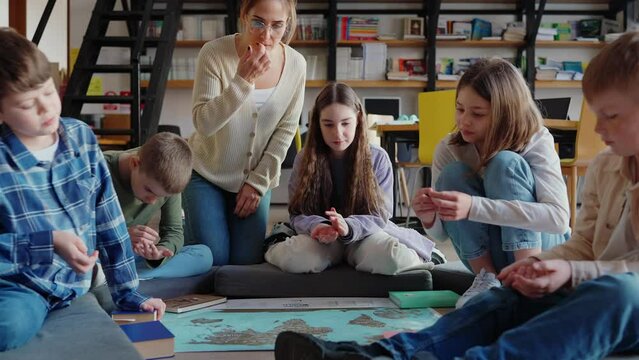 Positive children and teacher holding coins and looking at world map on the floor in library 