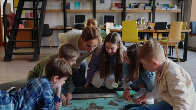Happy children and their teacher look at map of world sitting on floor in library 