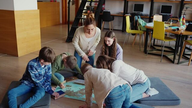 Confident children and their teacher look at map of world sitting on floor in library 