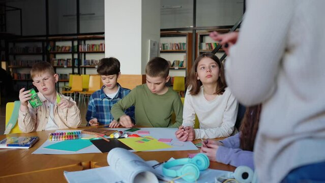 Pensive children listen to teacher before playing crocodile game at drawing table at library