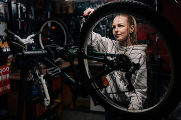 Fototapeta na wymiar Attractive skilled cycling repairman female repairing and fixing mountain bicycle standing on bike rack in repair workshop with dark interior. Concept of professional maintenance of bicycle transport.