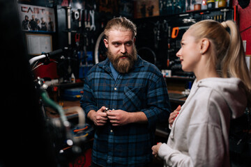 Fototapeta na wymiar Professional fashion cycling repairman with beard communicating with pretty blonde female client, talking problem of bicycle, detected during diagnostics in repair shop with dark interior.