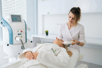 Obraz premium Portrait of female beautician doing hydro peeling procedure for woman in white bathrobe lying on medical couch in cosmetology clinic. Lady receiving stimulating electric facial treatment.