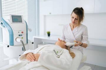 Portrait of female beautician doing hydro peeling procedure for woman in white bathrobe lying on medical couch in cosmetology clinic. Lady receiving stimulating electric facial treatment.