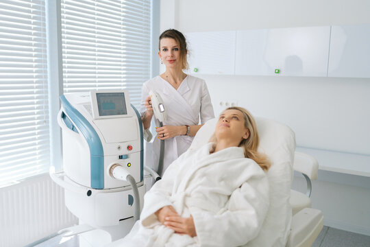 Portrait of female beautician specialist standing posing with device for photo rejuvenation in cosmetology clinic, looking at camera. Woman client receiving stimulating electric facial treatment.