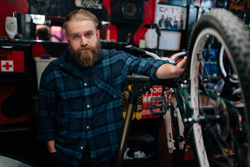 Fototapeta na wymiar Portrait of serious bearded cycling mechanic male standing by bicycle in repair bike workshop with dark interior, looking at camera. Concept of professional repair and maintenance of bicycle transport