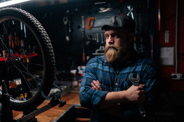 Portrait of stylish bearded repairman in cap holding wrench standing with crossed arms by bicycle in repair workshop with dark interior, looking away. Concept of professional bicycle maintenance.