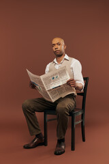 inclusion, bold african american man with myasthenia gravis syndrome, sitting on chair and reading newspaper, dark skinned black man with chronic disease on brown background