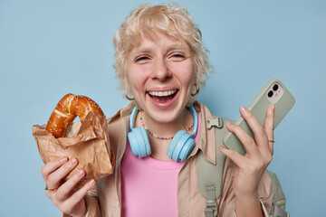 Happy blonde woman exudes zest for life poses with delicious bun in one hand and modern smartphone...