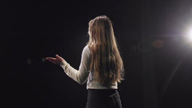 Back view of ecstatic caucasian woman clapping hands while standing on black stage with spotlights. Business motivator in casual attire cheering up audience for career growth and success.