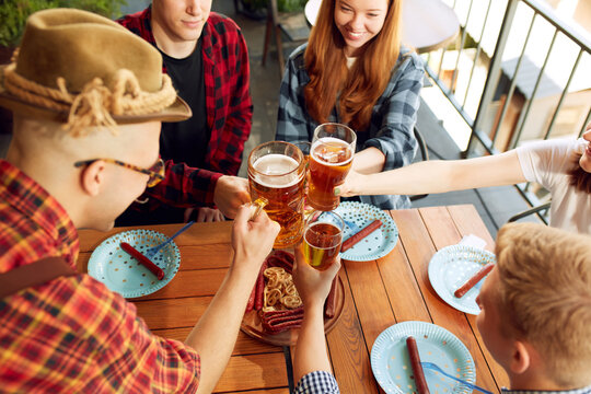 Tio view image of young people, friends cheerfully spending time together at cafe, pub, clinking glasses with lager beer. Concept of oktoberfest, traditional taste, friendship, leisure time, enjoyment