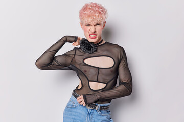 Seductive gay man has makeup with pink hair frowns face clenches teeth being dissatisfied with something wears black jumper and jeans looks negatively at camera isolated over white background