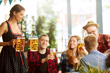 Beautiful young woman, waitress serving big mugs with lager beer to group of young people, friends meeting together at pub. Oktoberfest, traditional taste, friendship, leisure time, enjoyment concept
