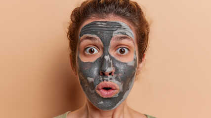 Headshot of young woman indulges in pampering skincare routine stares impressed has mouth opened applies beauty clay mask to rejuvenate skin isolated over brown background. Wellness concept.