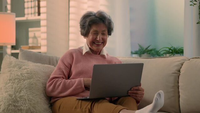 Caucasian happy cheerful old woman senior lady mature retirement granny elderly female at couch home talk laptop video chat call talking to computer web camera conference with grandchildren children