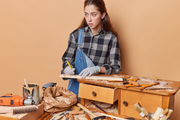 Female carpenter passionately marks something on wooden plank with marker dessed in workwear captures essence of dedication to joinery surrounded by different tools repairs old furniture. Carpentry