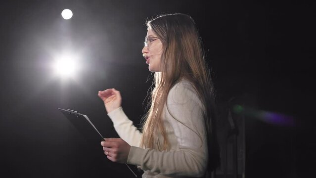 Side view of caucasian woman in eyeglasses talking and gesturing on stage during business performance. Motivational speaker with headset and clipboard explaining listeners strategies for success.