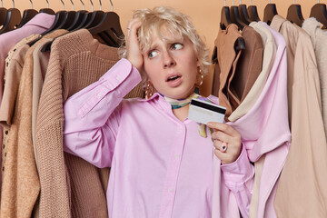 Photo of European confused lady standing near clothing rail choosing new stylish outfit holding...